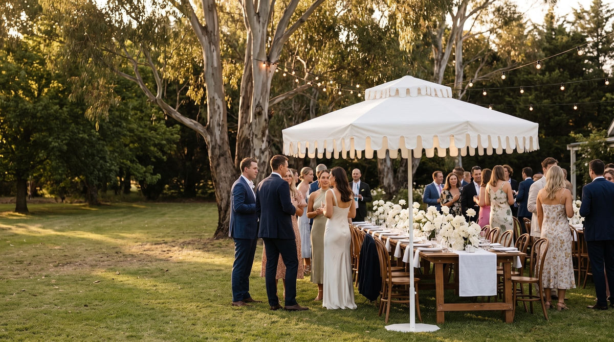 A long wedding table under an Ivory Mist umbrella at golden hour, with guests, gum trees and festoon lights